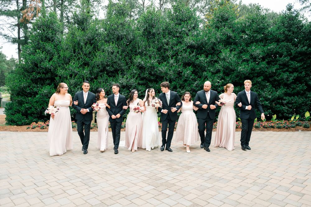 Wedding party walking together outdoors, bridesmaids in blush pink gowns and groomsmen in black suits, with bride and groom centered.