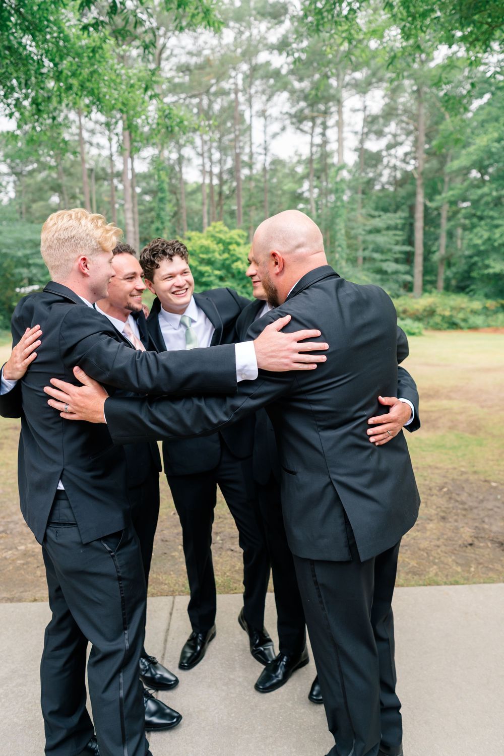 Groomsmen in black suits sharing a group hug outdoors surrounded by tall pine trees at a wedding.