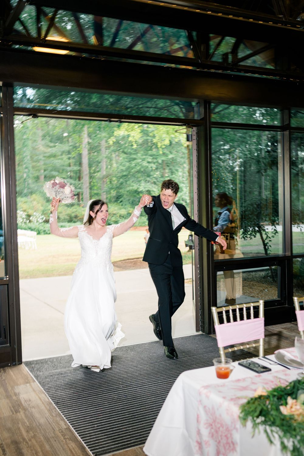 Bride and groom make joyful grand entrance at wedding reception, bride holding bouquet aloft in elegant white gown.