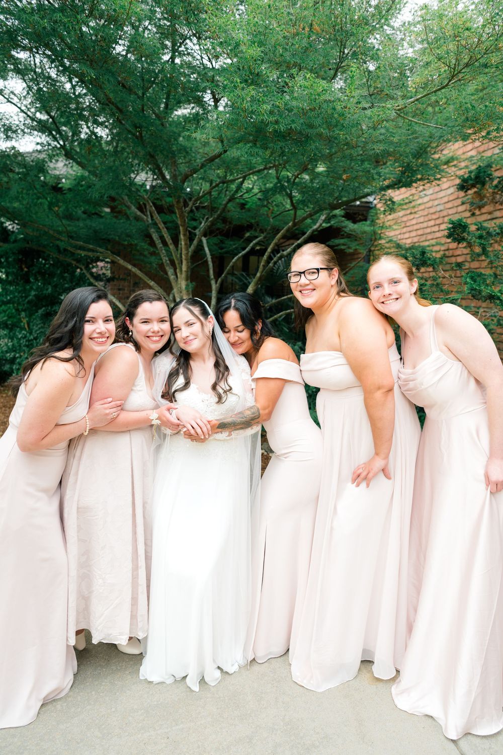 Bride in white gown poses with five bridesmaids in blush pink dresses, laughing together outdoors near brick wall.