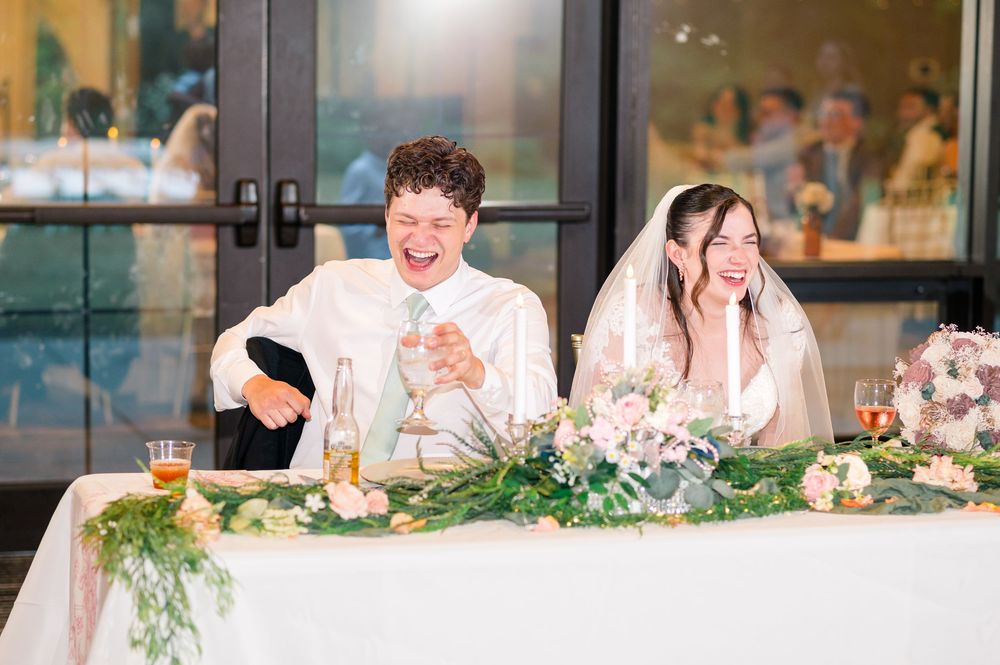 Bride and groom laughing joyfully at sweetheart table during wedding reception with floral centerpiece.