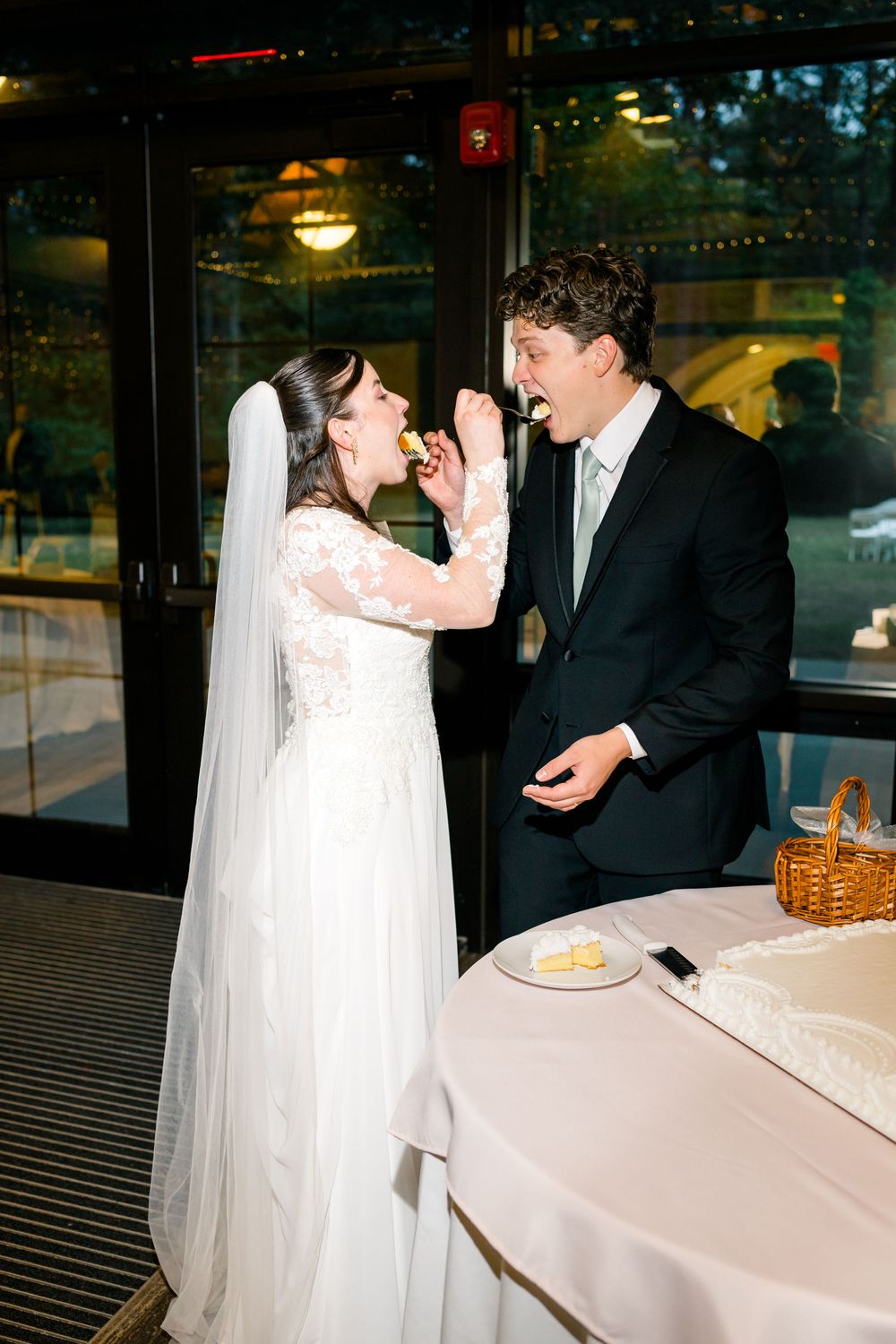 Bride feeds groom wedding cake during evening reception near entrance doors of elegant venue.