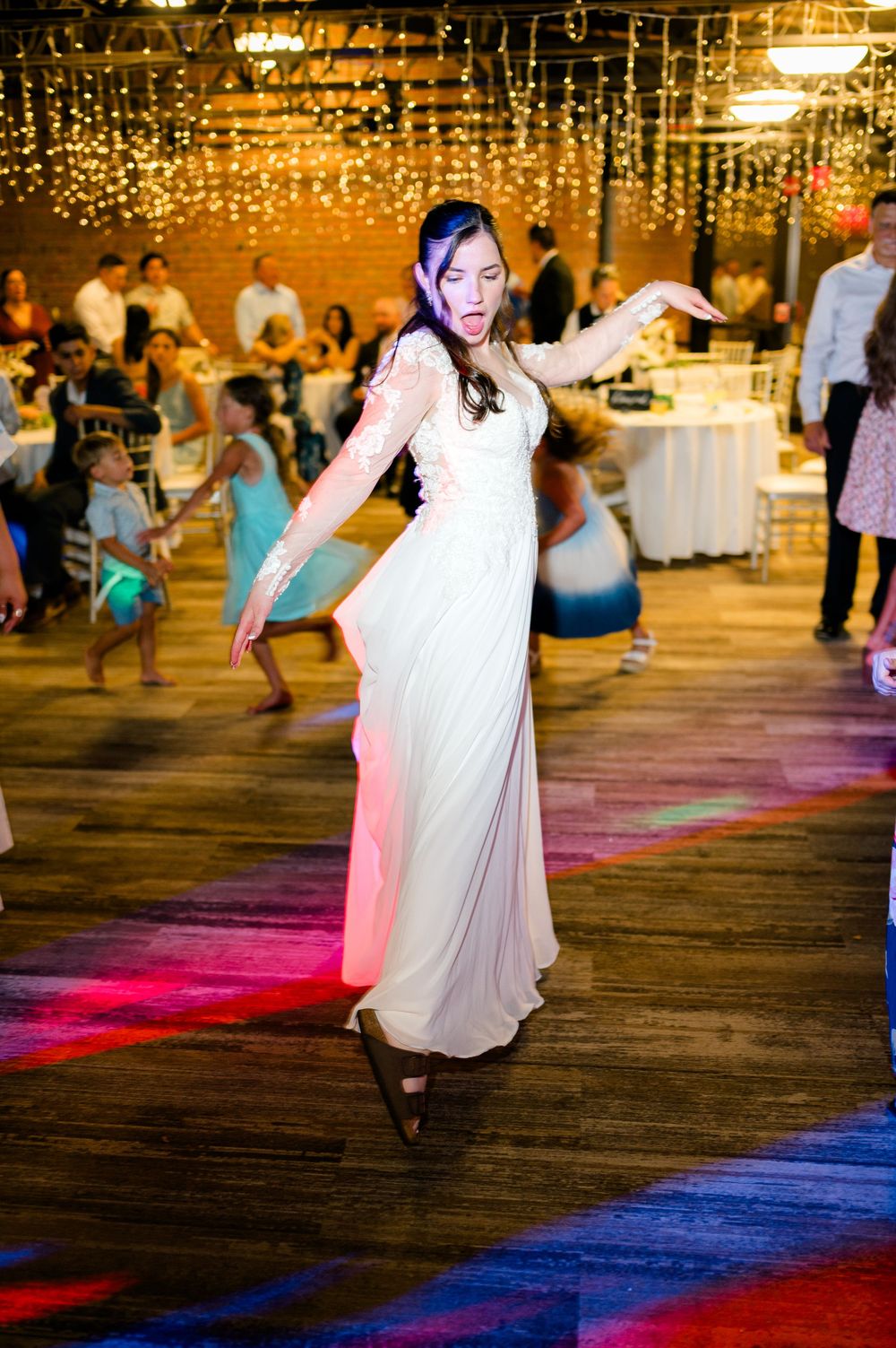 Bride in white lace gown dancing joyfully on colorful lit dance floor at elegant wedding reception.