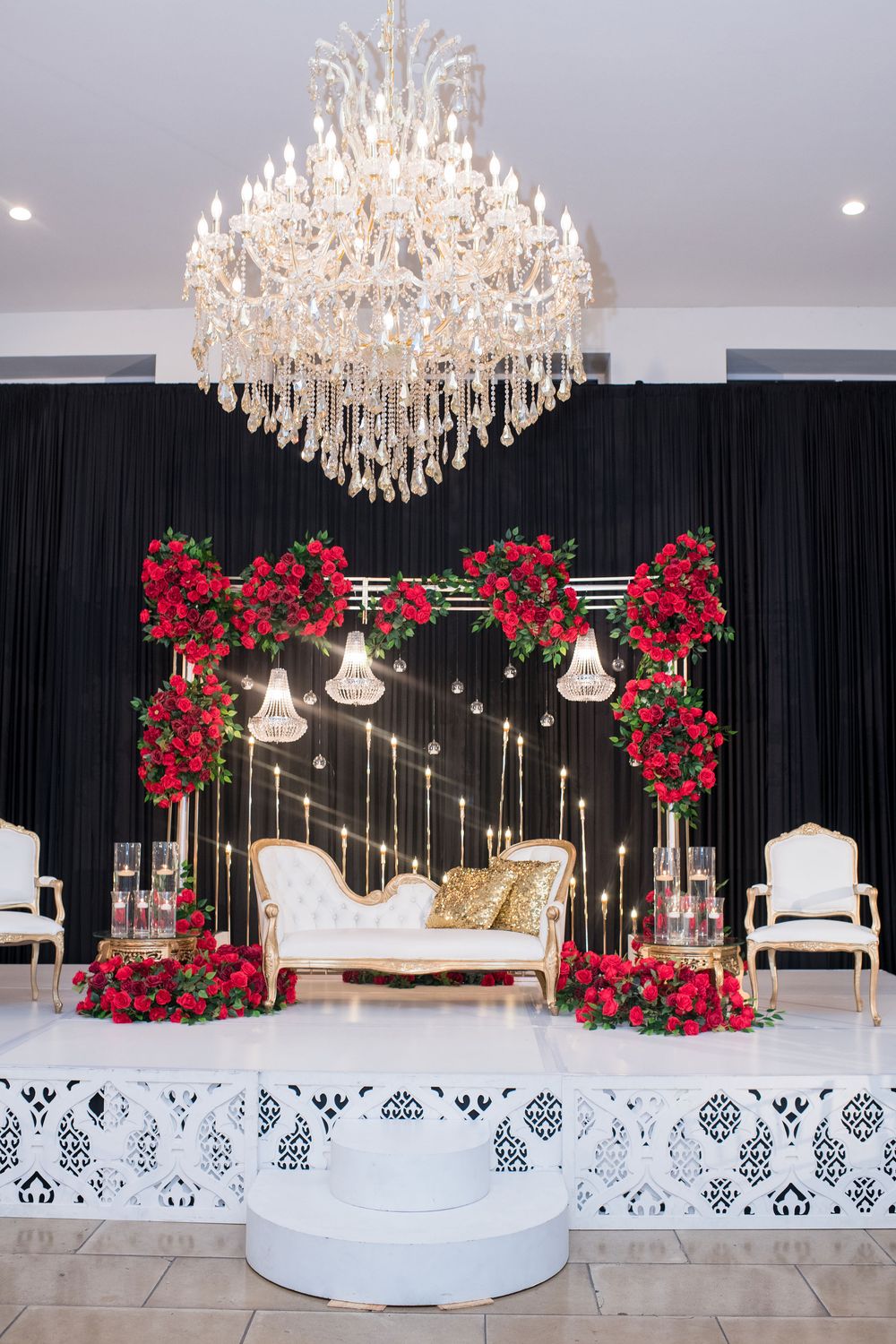 Elegant wedding stage with white sofa, red rose arch, crystal chandelier, and black drape backdrop.
