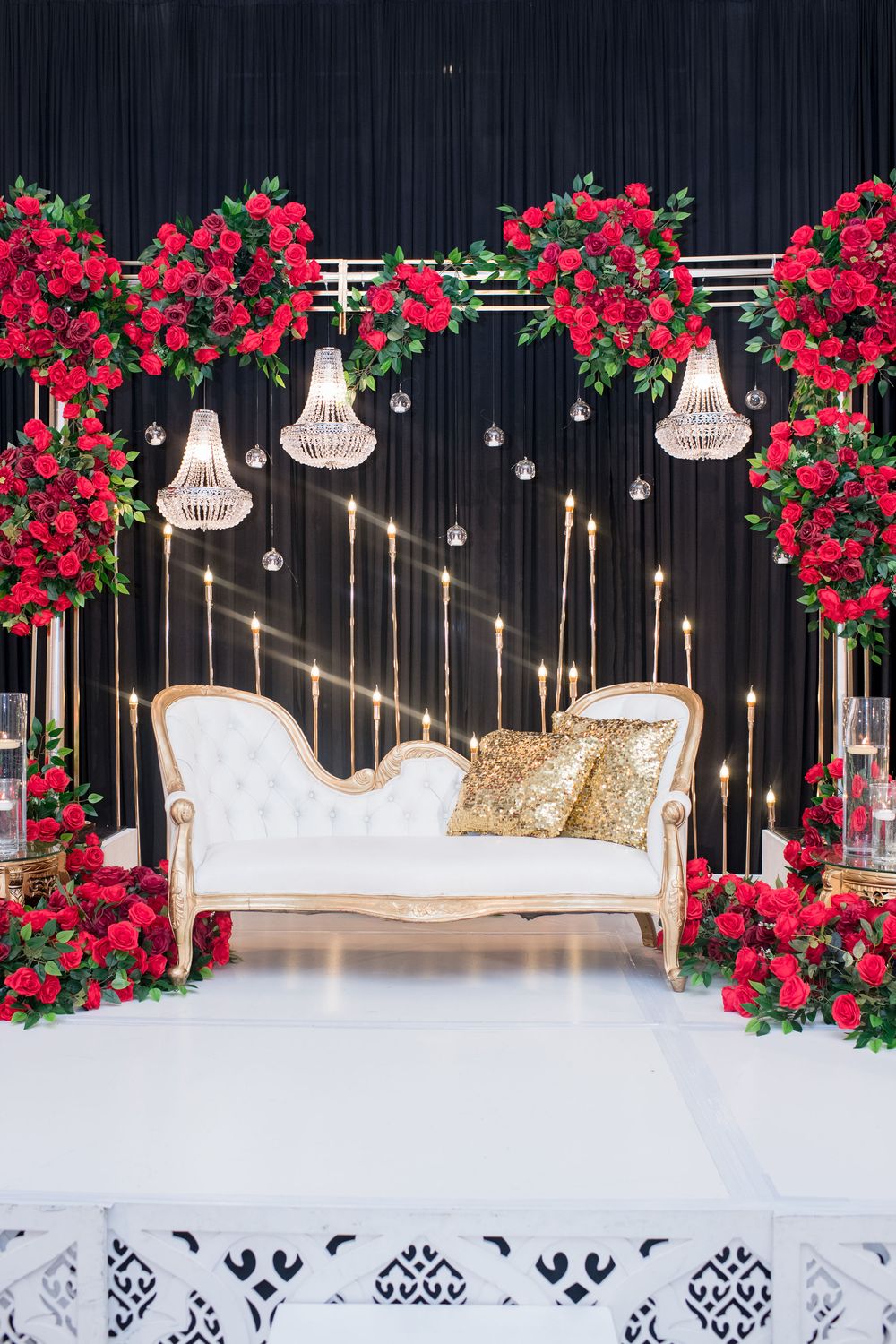 Elegant wedding stage with white tufted sofa, red roses, crystal chandeliers, and gold pillows on black backdrop.
