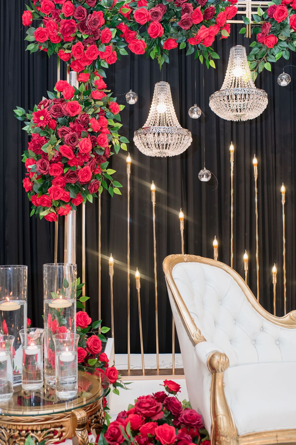 Elegant wedding decor with red roses, crystal chandeliers, white throne chair, and gold candle stands on black backdrop.