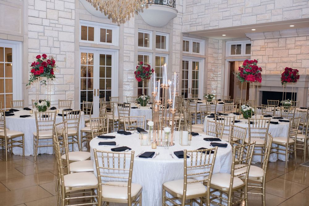Elegant banquet hall with gold chiavari chairs, white round tables, red floral centerpieces, and crystal chandelier.