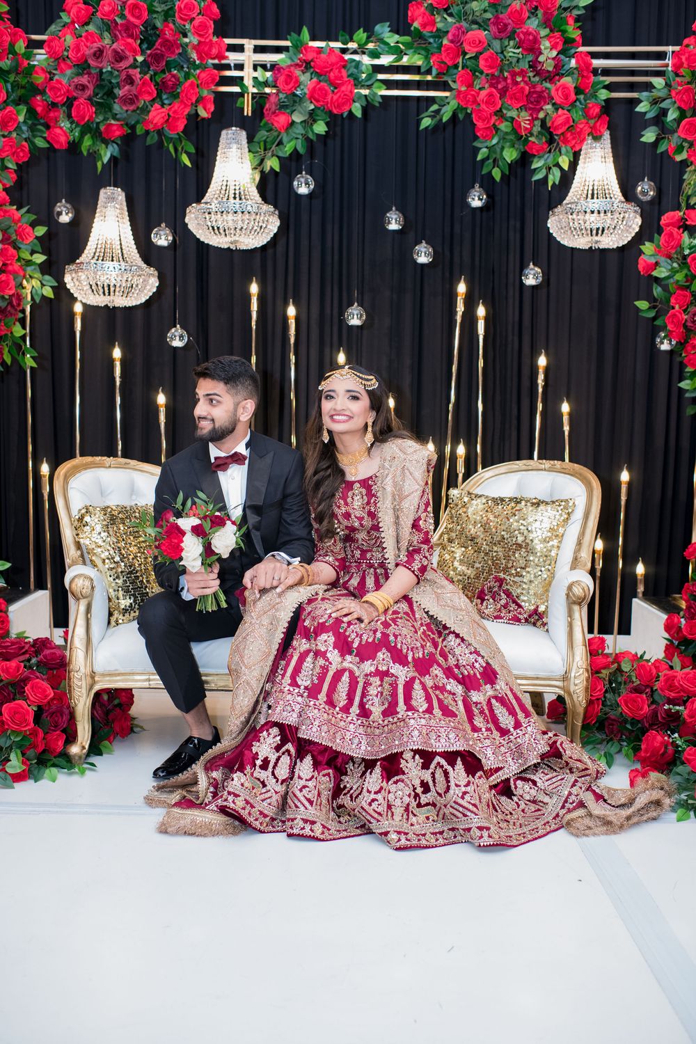 South Asian bride and groom in traditional red and gold wedding attire seated on ornate chairs with floral decor.
