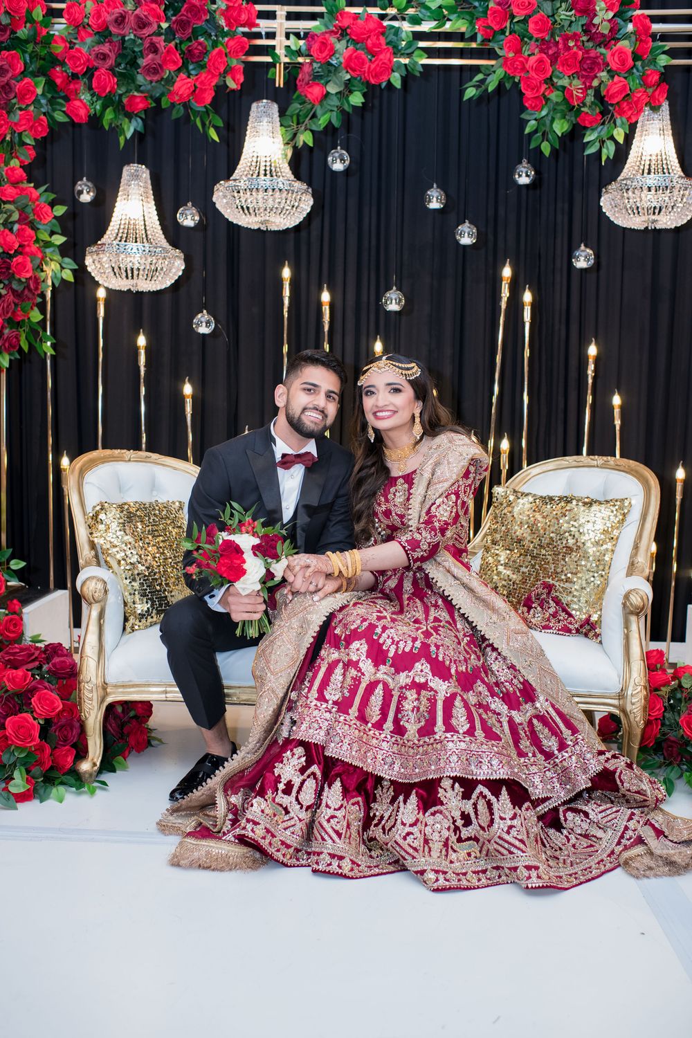 South Asian bride in red lehenga and groom in black tuxedo pose on ornate chairs with crystal chandeliers and red roses.