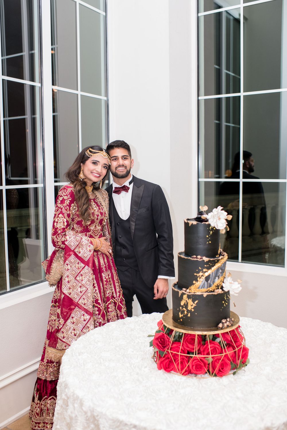 South Asian bride in red lehenga and groom in black tuxedo pose beside elegant black and gold wedding cake.