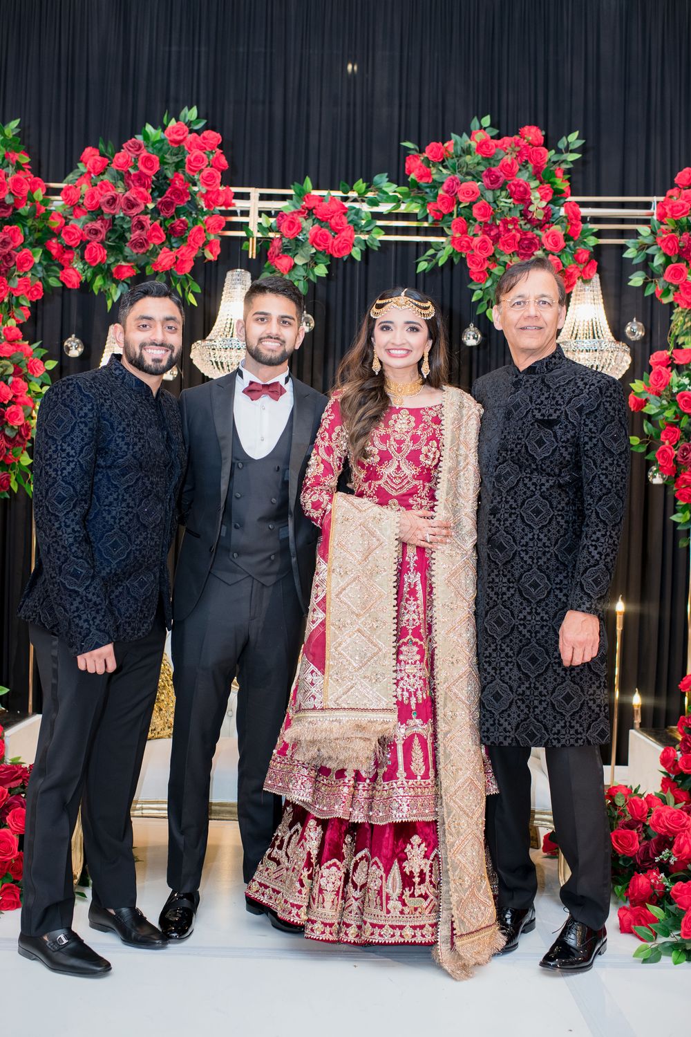 South Asian bride in red bridal lehenga poses with three men in formal wear against red rose floral backdrop.