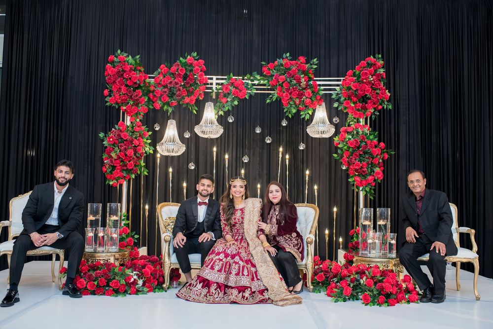 South Asian wedding couple in red and black attire seated at ornate floral backdrop with rose arrangements and candles.