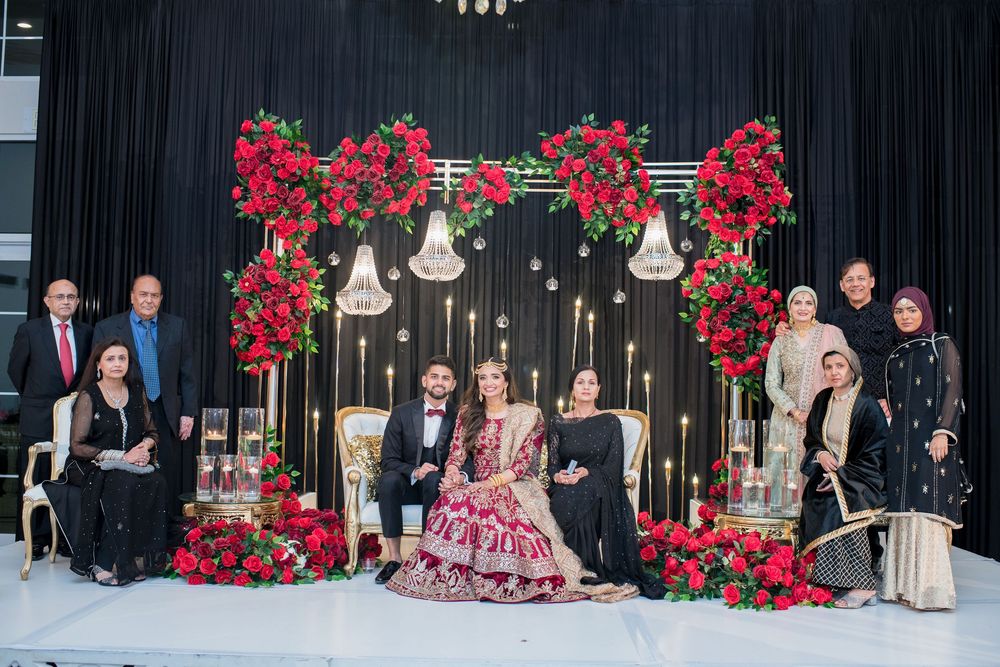 South Asian wedding couple in traditional attire seated with family before red rose floral backdrop with gold candelabras.
