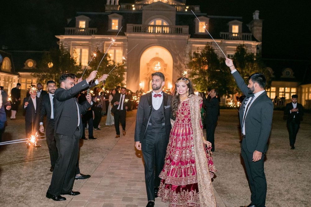 Bride in red lehenga and groom in gray suit walk through sparkler send-off at elegant mansion wedding venue at night.