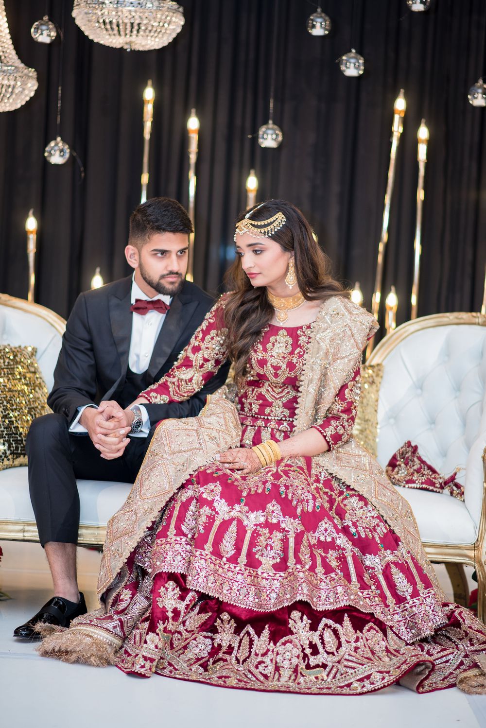Bride in red and gold lehenga with groom in black tuxedo seated on white sofa at elegant wedding reception.