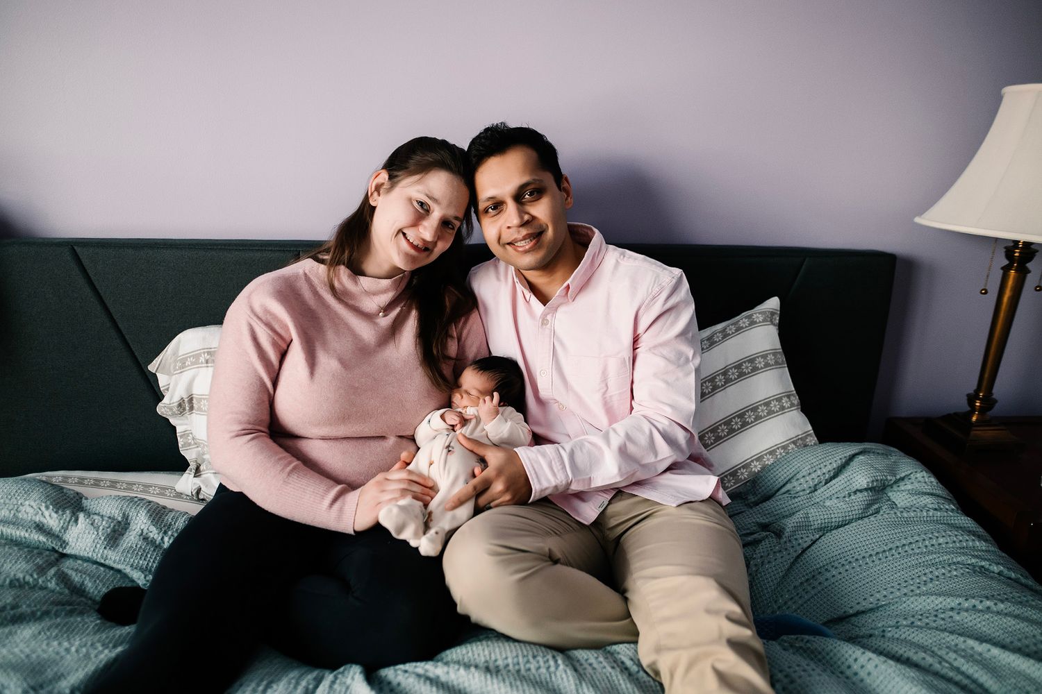 Happy family with newborn baby sitting together on a bed, smiling warmly in a cozy bedroom setting.