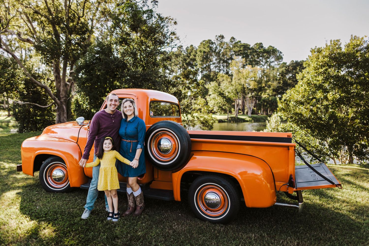 Family of three posing beside a vintage orange pickup truck on green grass surrounded by trees.