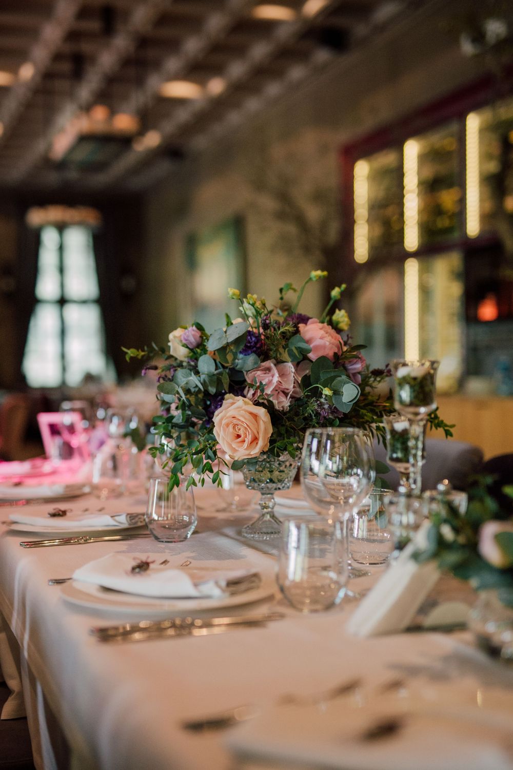 Elegant wedding reception table with blush pink roses, purple flowers, and greenery centerpiece, white linens, and crystal glassware.