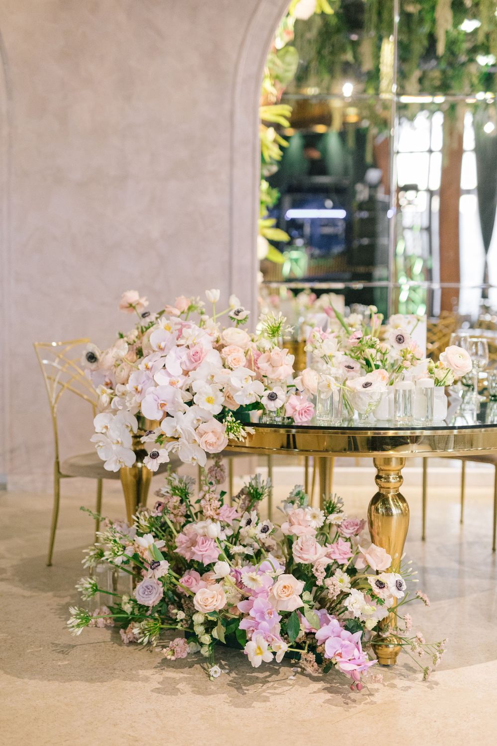 Elegant wedding table with lush pink and white floral arrangements on a gold base, set in a softly lit venue.