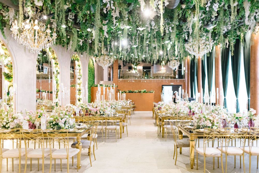 Elegant wedding reception hall with gold chairs, floral centerpieces, and lush greenery hanging from crystal chandeliers.