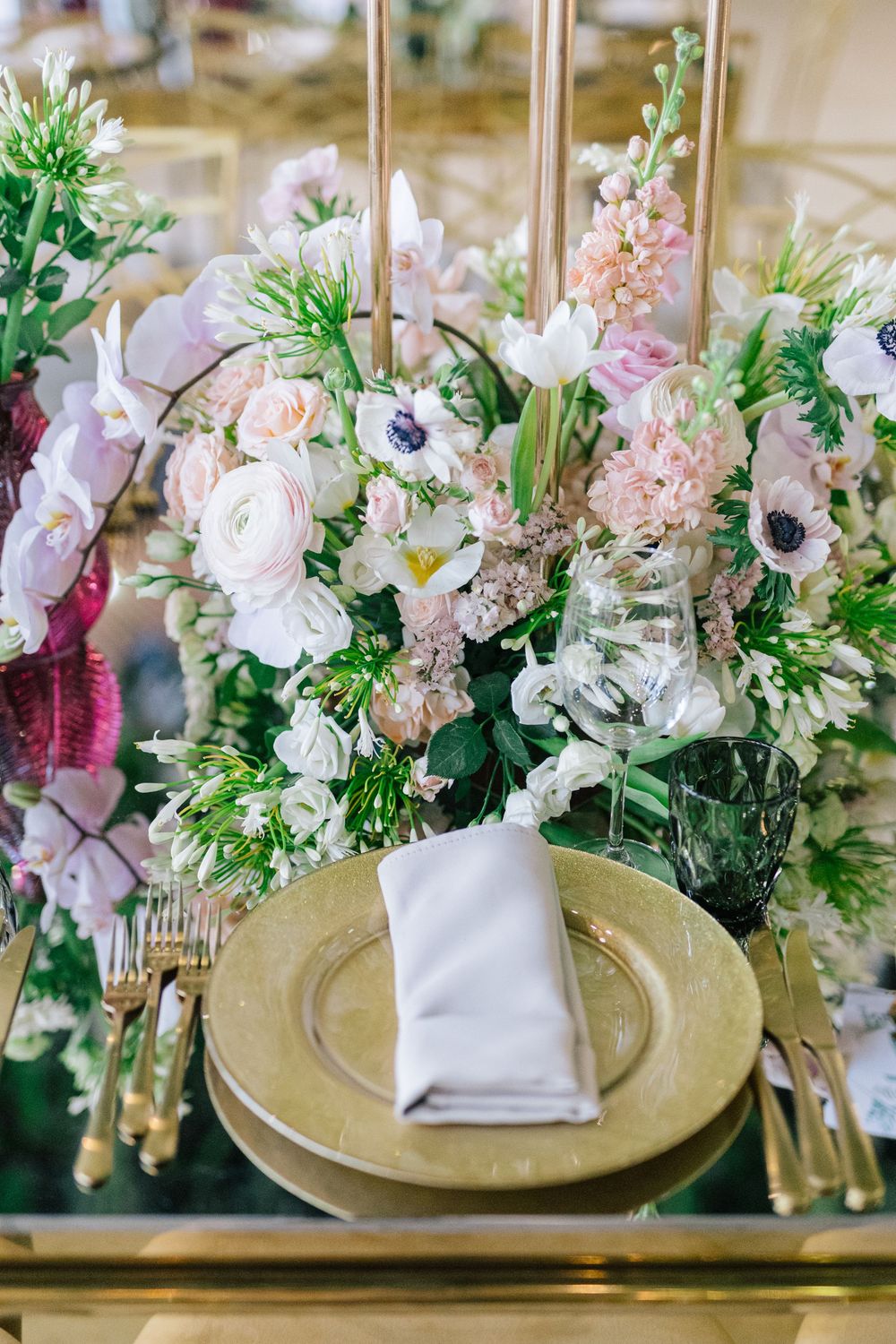 Elegant wedding table setting with gold chargers, white napkin, and lush floral centerpiece in blush and white tones.
