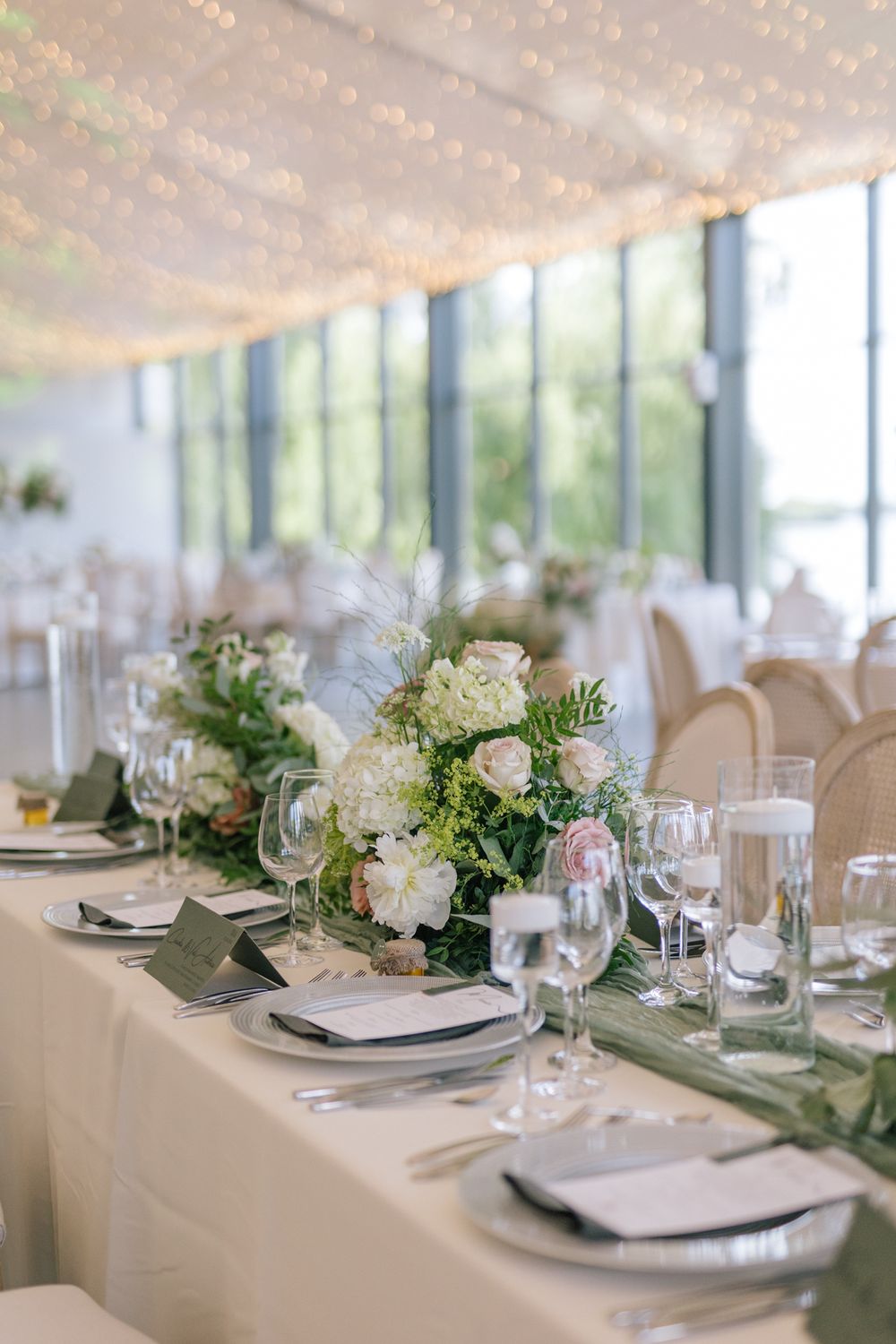 Elegant wedding reception table with floral centerpiece, white and pink blooms, green runner, crystal glasses, and fairy lights.