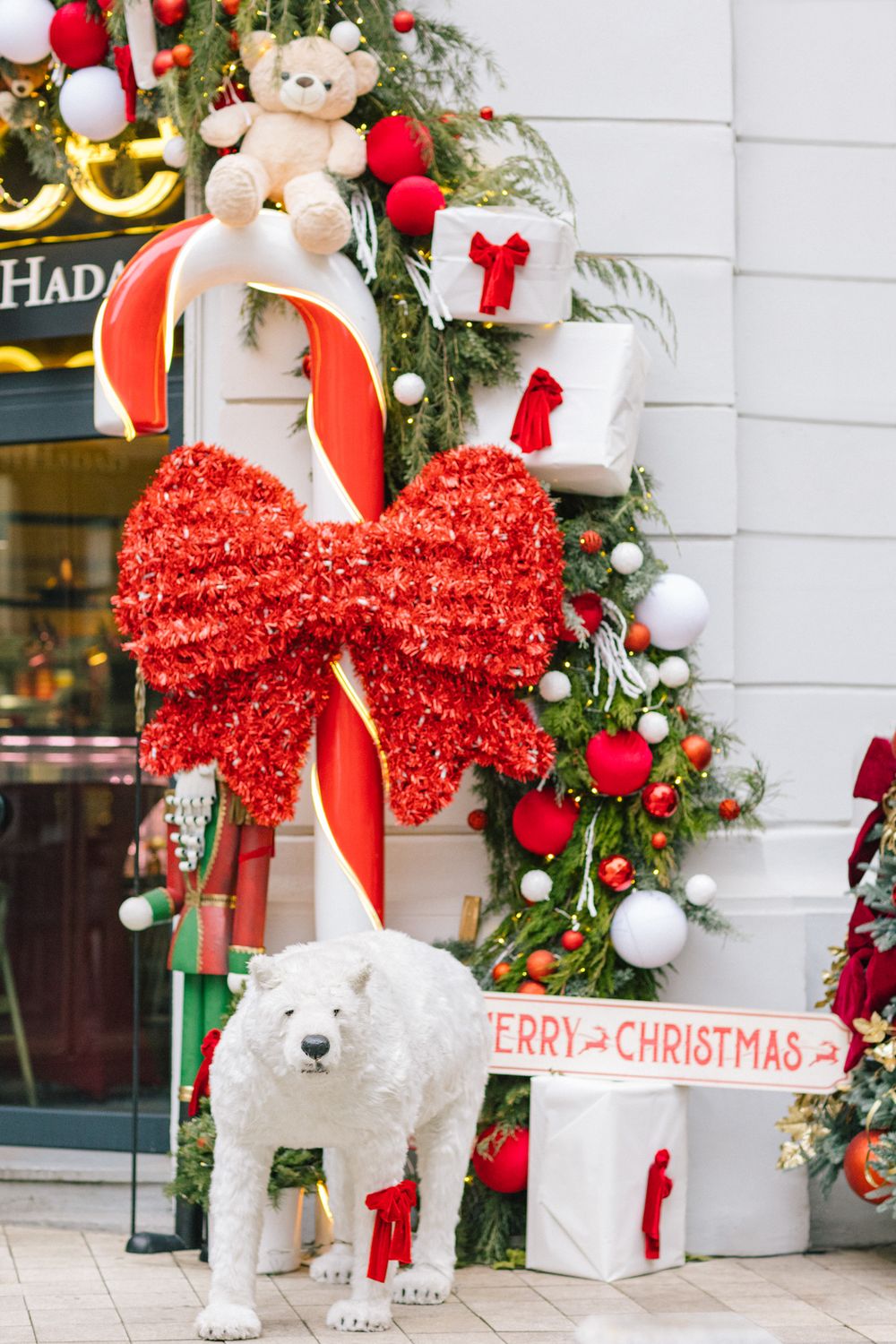 White polar bear statue with Merry Christmas sign amid festive red bow, candy cane, and holiday decorations outside a store.