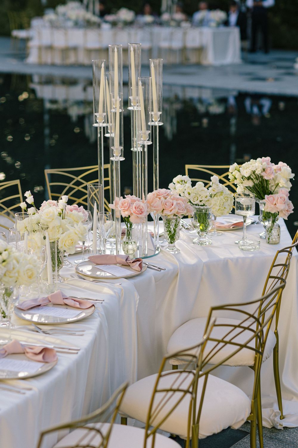 Elegant wedding reception table with gold chairs, crystal candelabras, and blush pink roses on white linen outdoors.