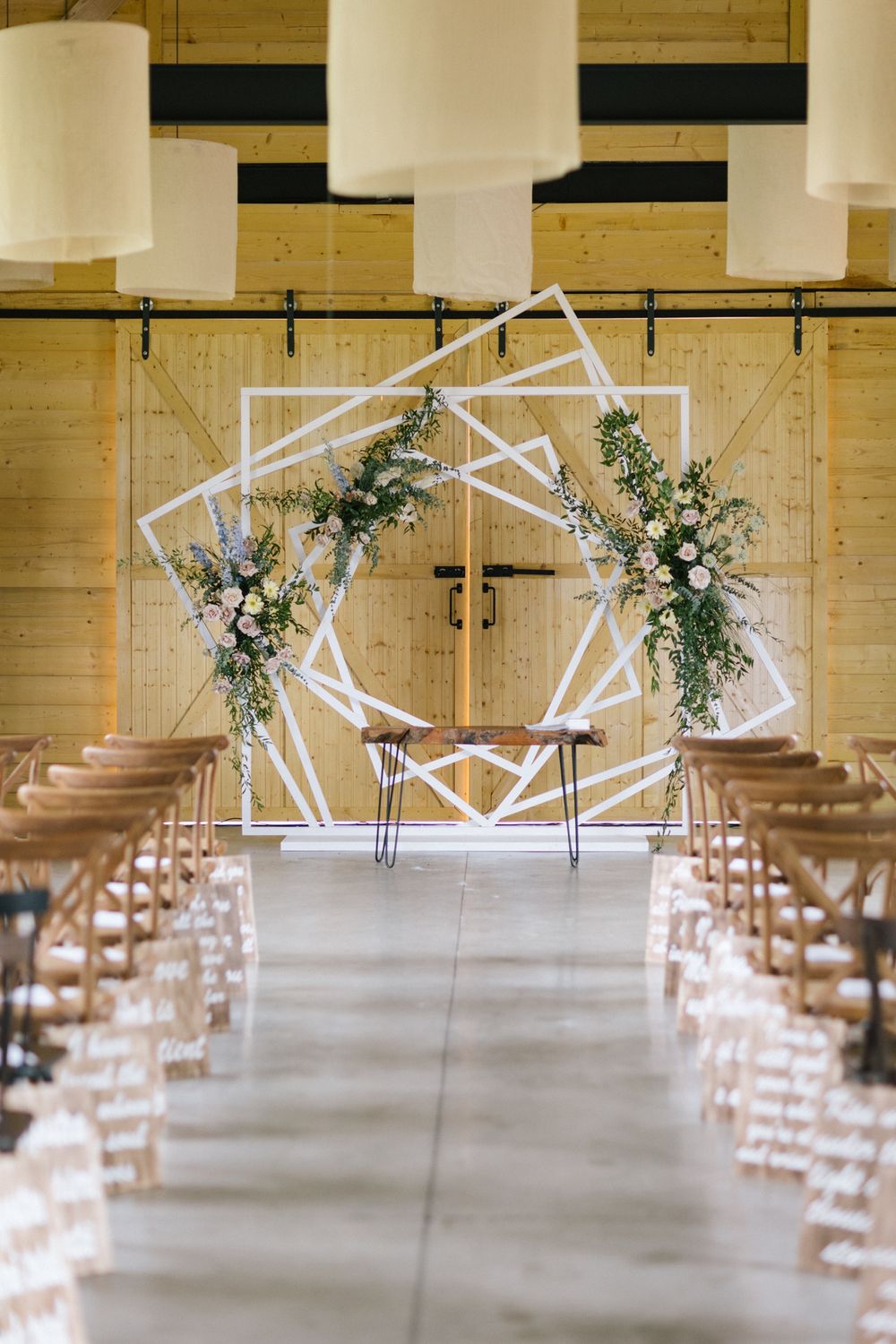 Elegant barn wedding ceremony with geometric white arch, floral arrangements, and rows of wooden chairs on concrete floor.
