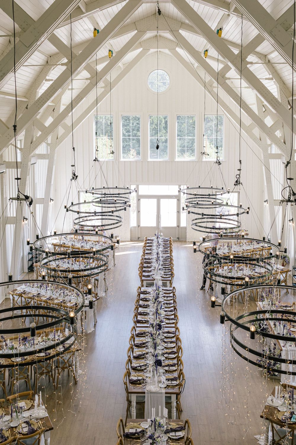Elegant white barn wedding venue with vaulted ceilings, circular chandeliers, and long reception tables set for a formal dinner.