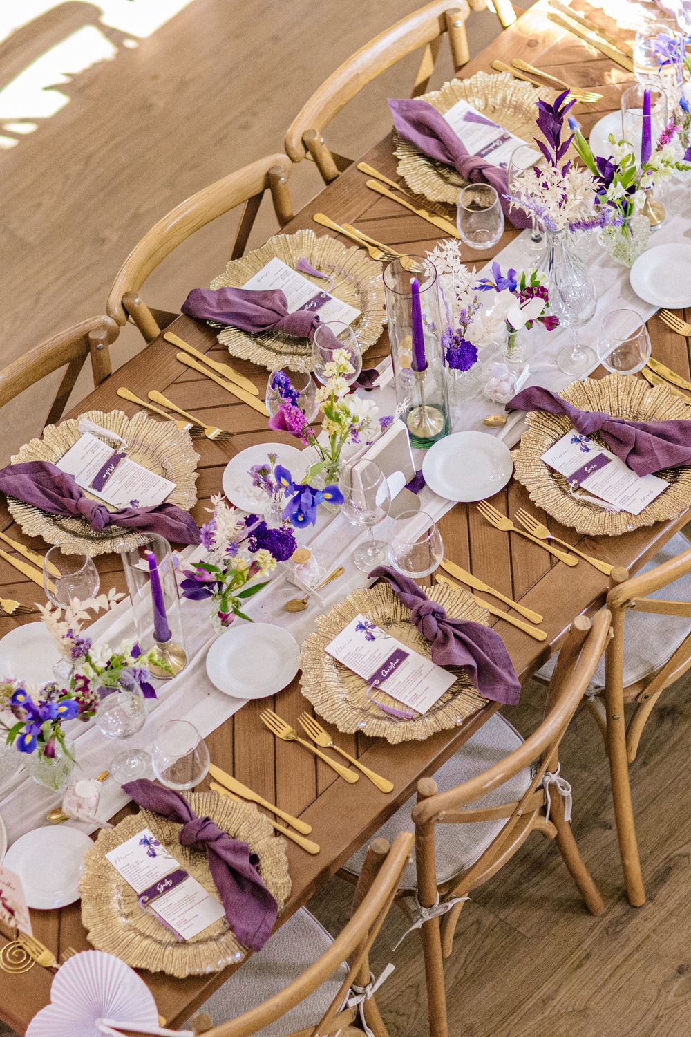 Elegant wedding table setting with purple napkins, gold cutlery, wicker chargers, and floral centerpieces on a wooden table.