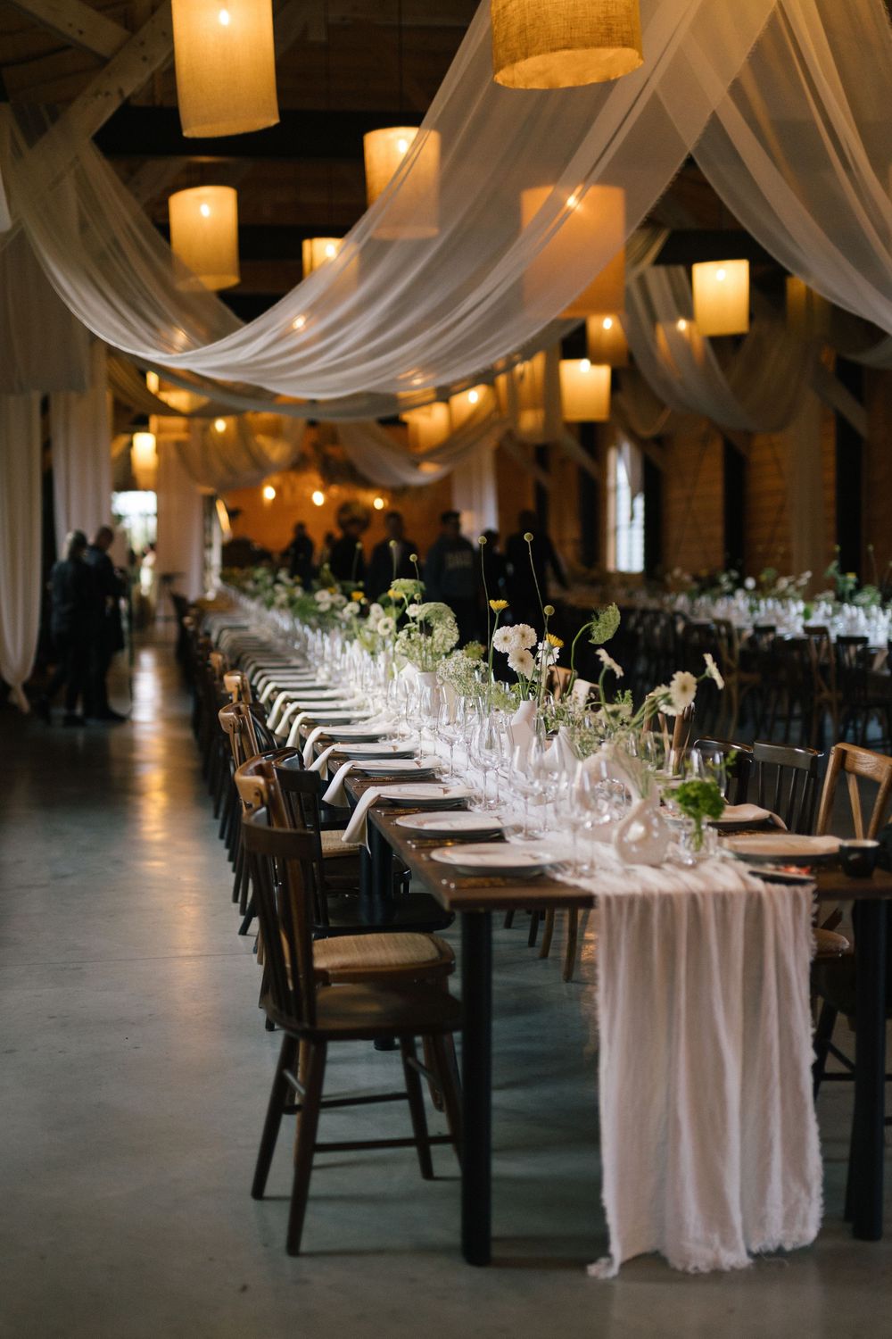 Elegant wedding reception hall with white draped ceilings, long wooden tables, floral centerpieces, and warm pendant lighting.
