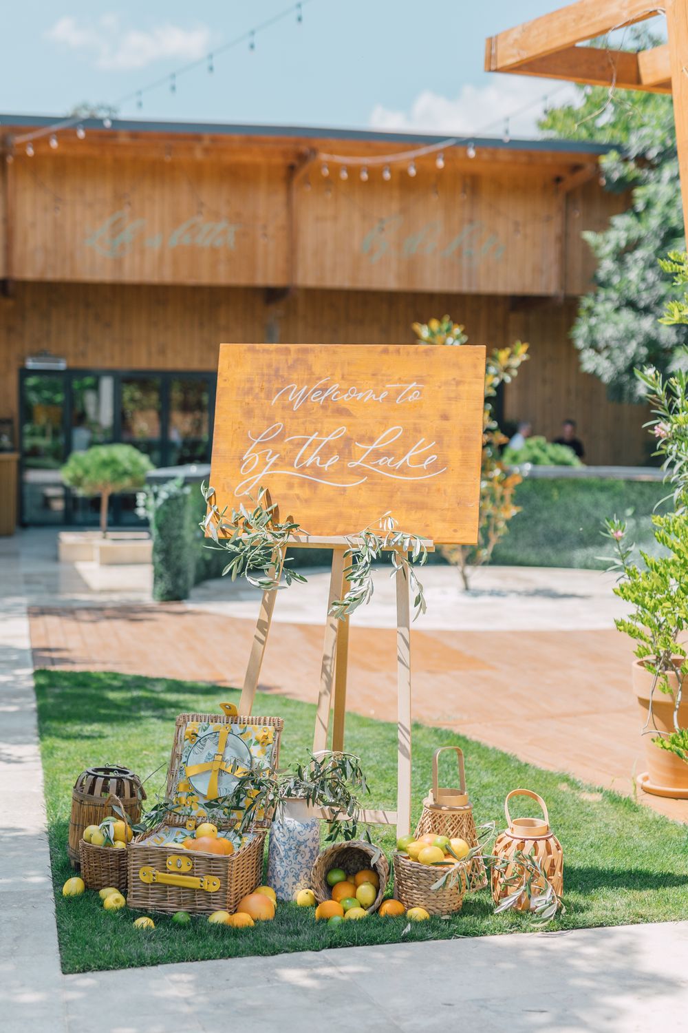 Outdoor wedding welcome sign reading By the Lake surrounded by wicker baskets and fresh lemons on green grass.