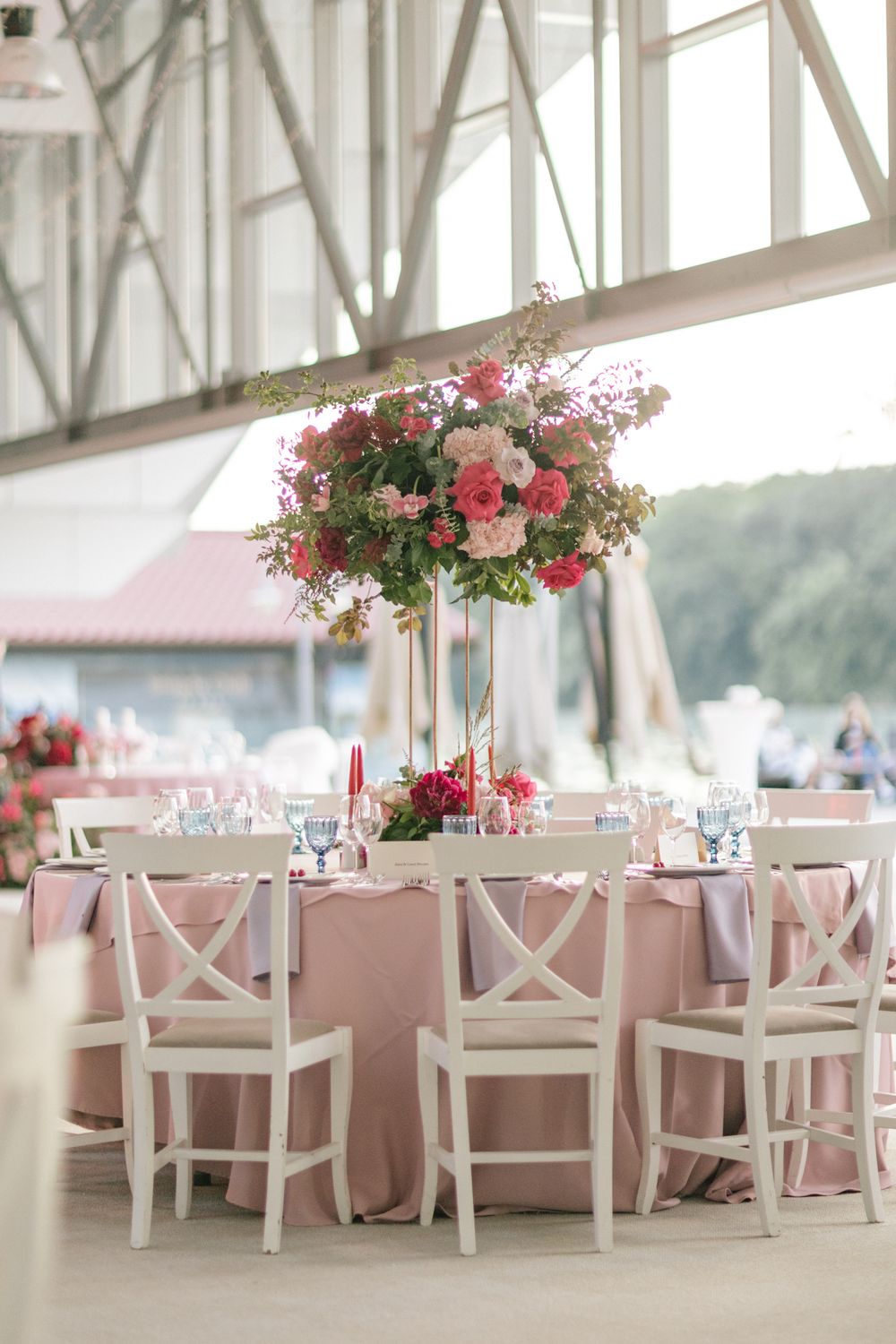 Elegant wedding reception table with pink tablecloth, white cross-back chairs, and tall floral centerpiece with pink blooms.