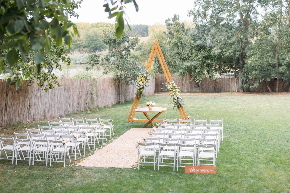 Outdoor wedding ceremony setup with white chairs, a triangular wooden arch adorned with flowers, on a lush green lawn.
