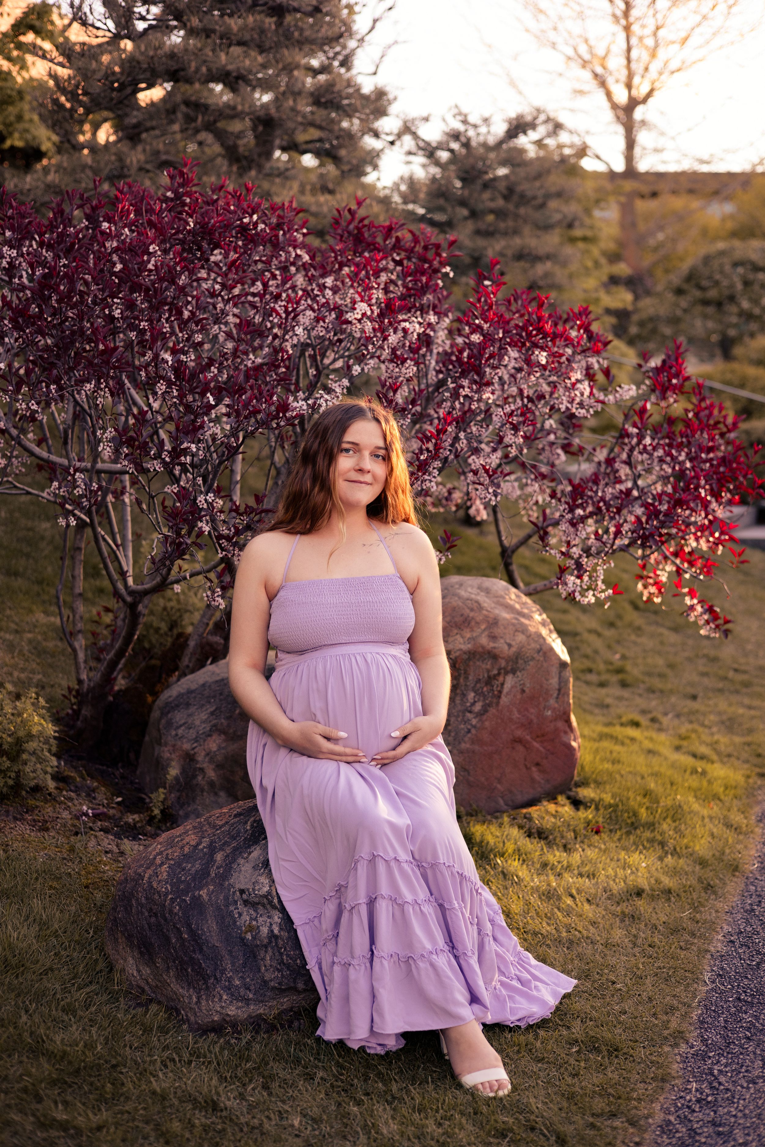 Pregnant woman in lavender maxi dress sitting on rocks surrounded by blooming spring trees at golden hour.