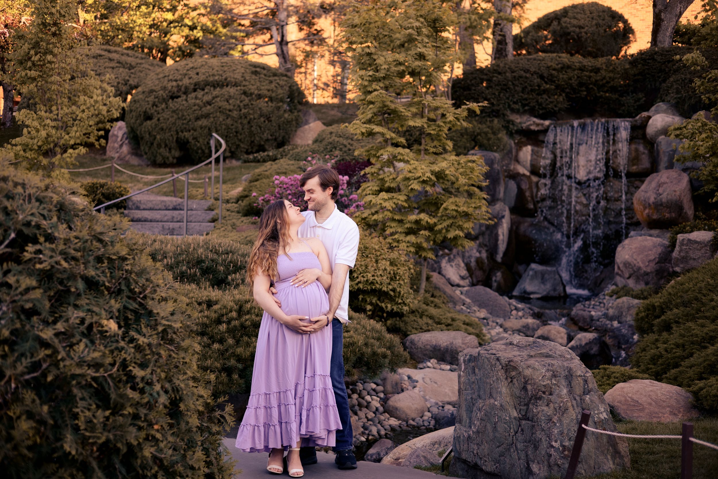 Pregnant couple embracing in a lush garden with a waterfall, woman in lavender dress, golden hour lighting.