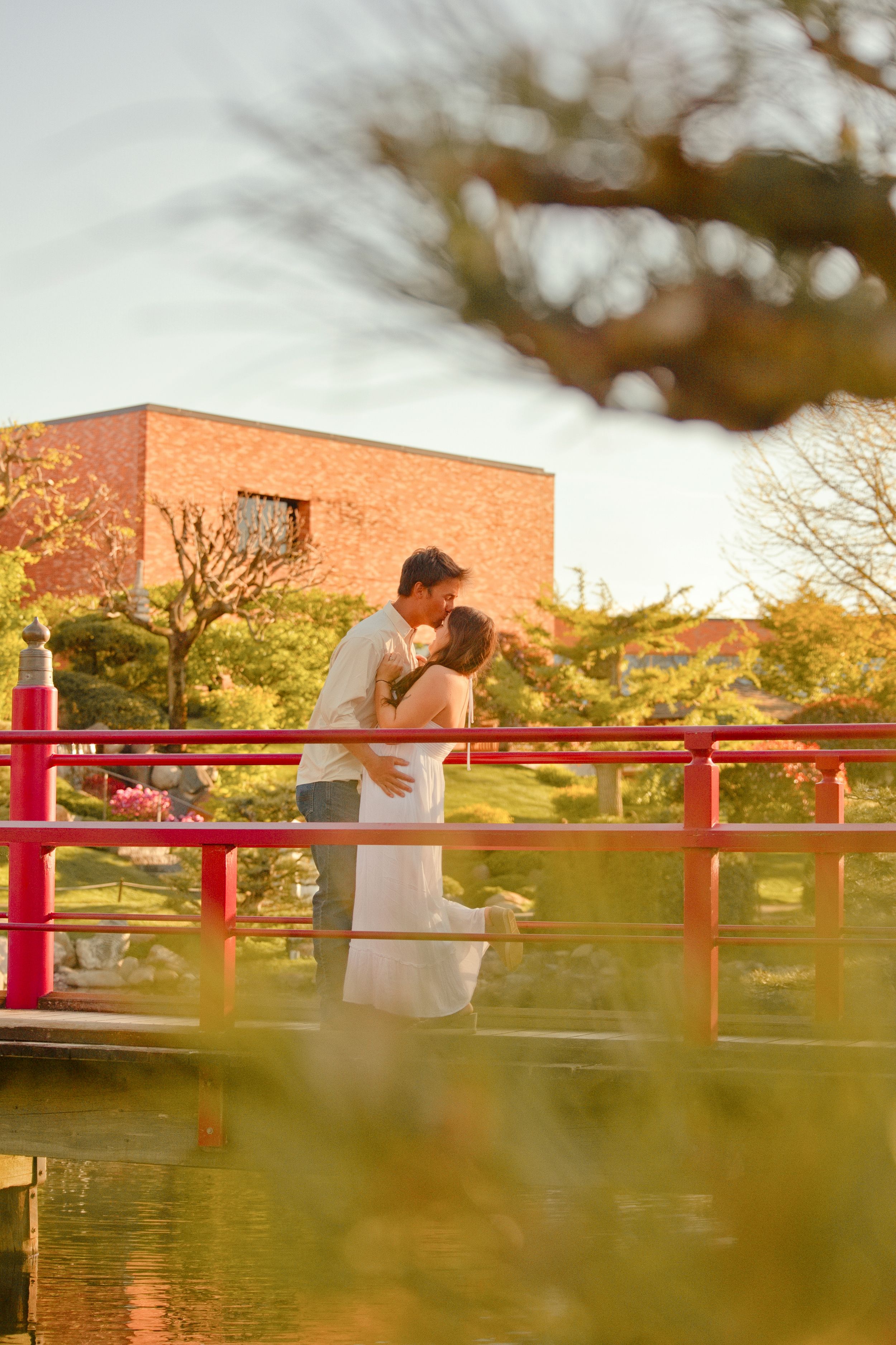 Couple sharing a romantic kiss on a red Japanese garden bridge, surrounded by lush greenery and golden sunset light.