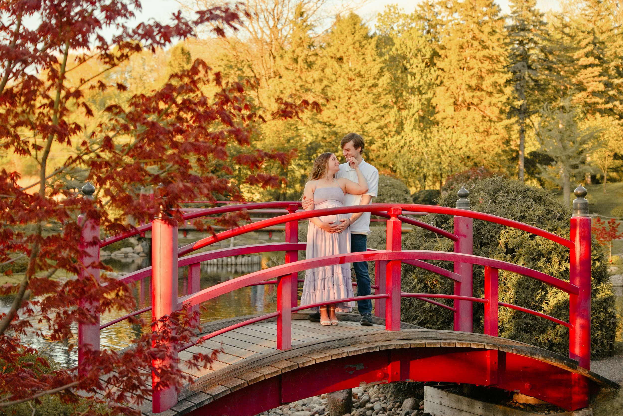 Couple embracing on a red arched bridge surrounded by vibrant autumn foliage in a Japanese garden.