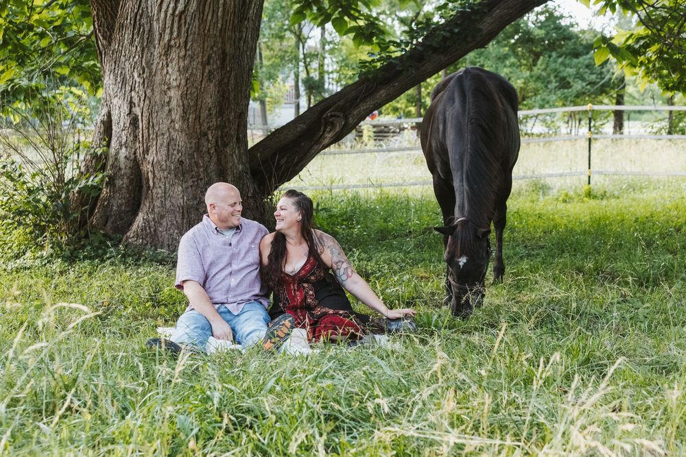 Couple sitting together in green grass under a large tree beside a dark horse in a sunny outdoor setting.