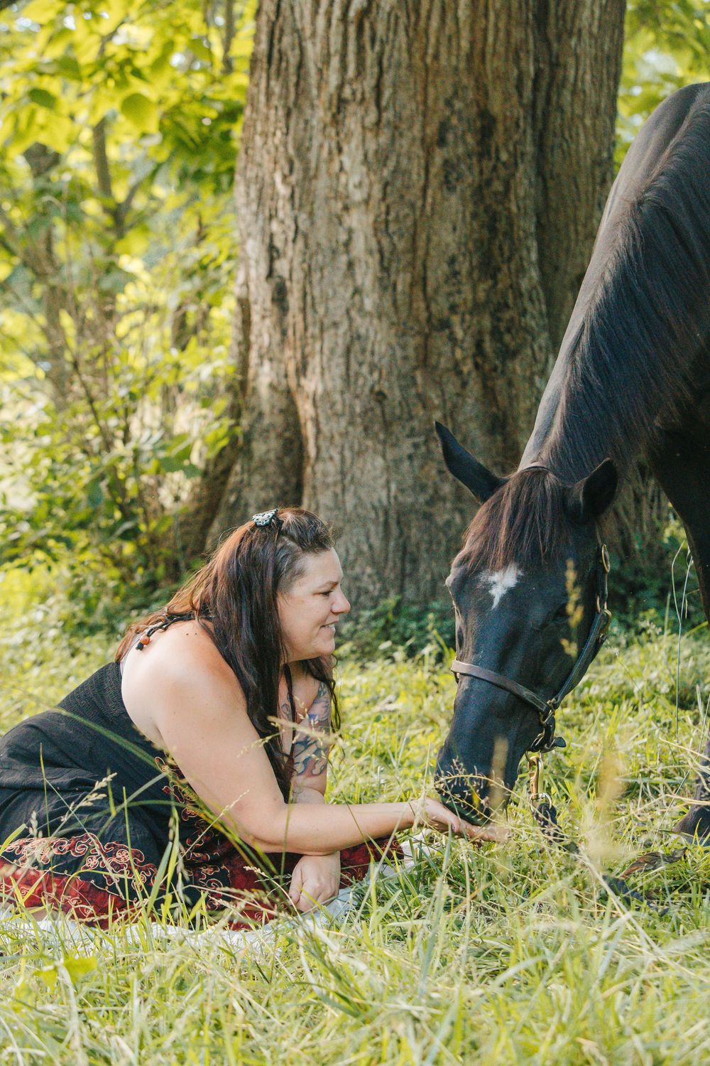 Woman smiling while feeding a dark horse by hand, sitting in green grass near a large tree outdoors.