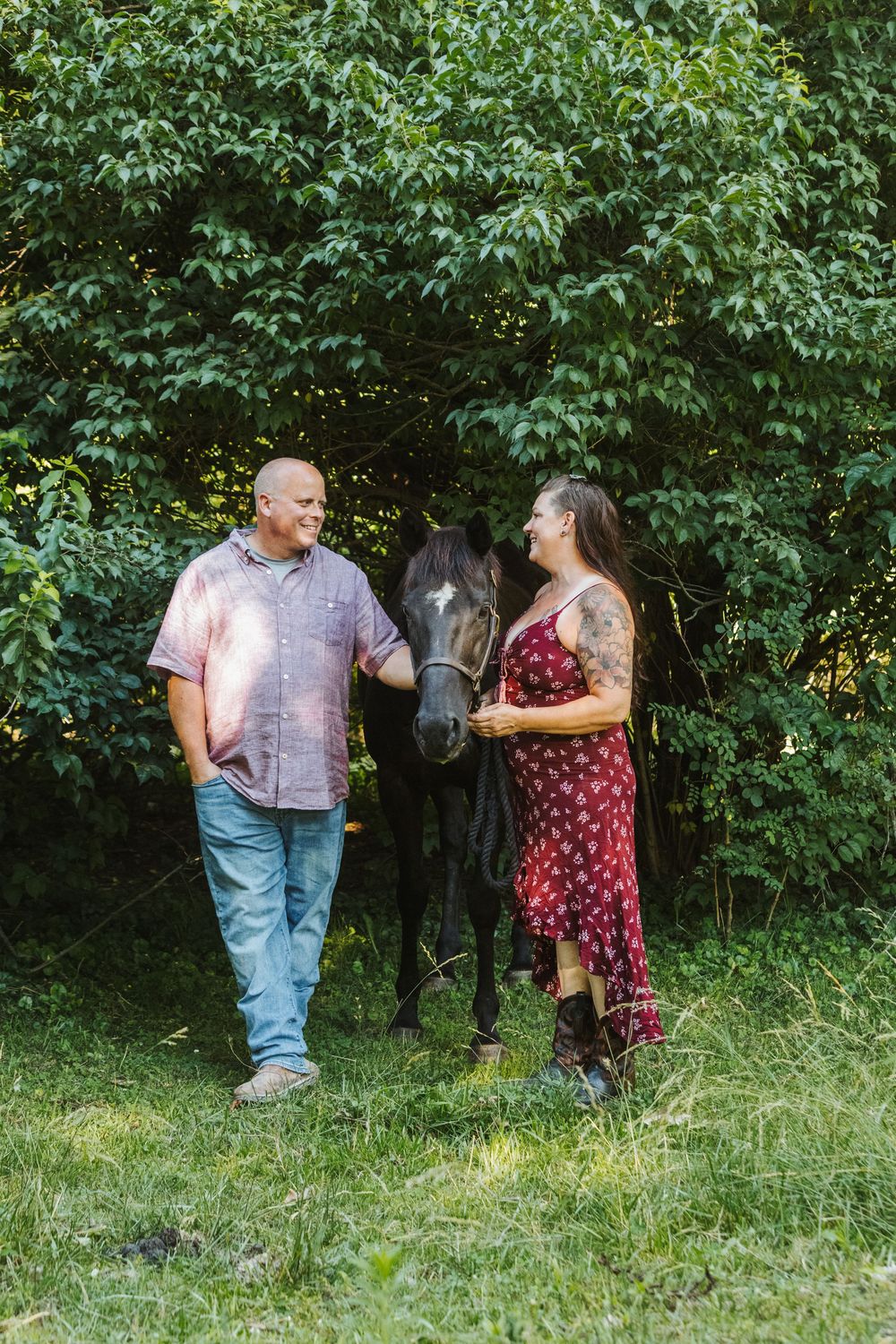 A couple smiles at each other beside a dark horse surrounded by lush green trees outdoors.