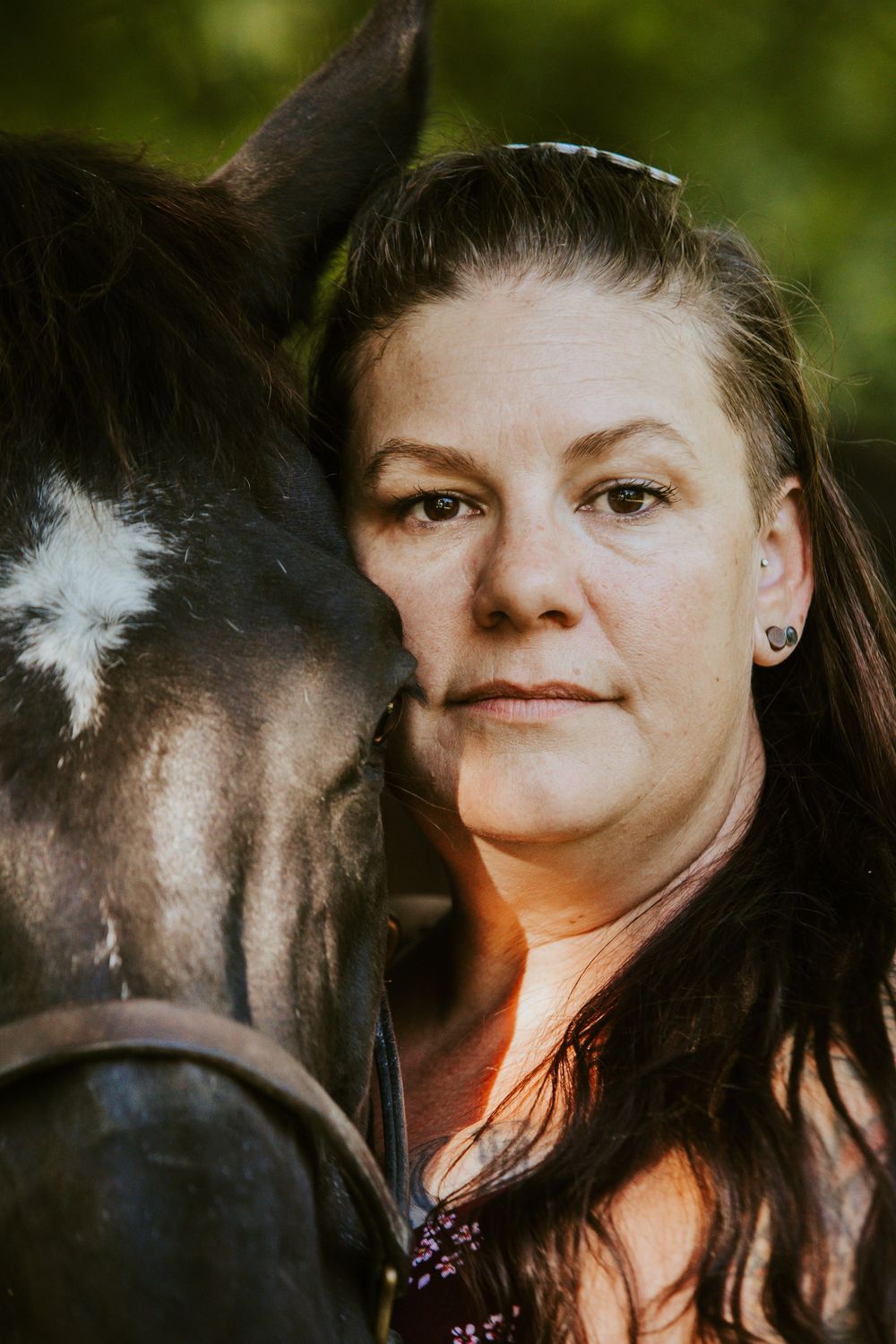 Woman with dark hair poses closely with a dark horse outdoors, sharing a serene moment in natural green surroundings.