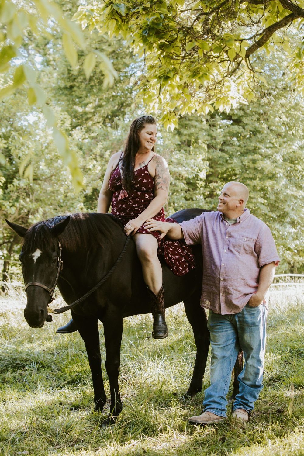 A woman in a floral dress sits on a black horse while a bald man in a pink shirt stands beside her outdoors.