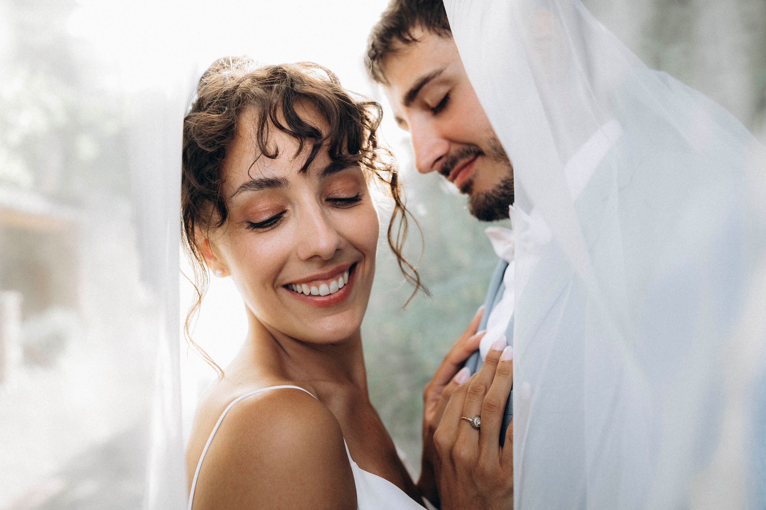 A joyful bride smiles under a white veil while a groom leans in close during a romantic wedding moment.