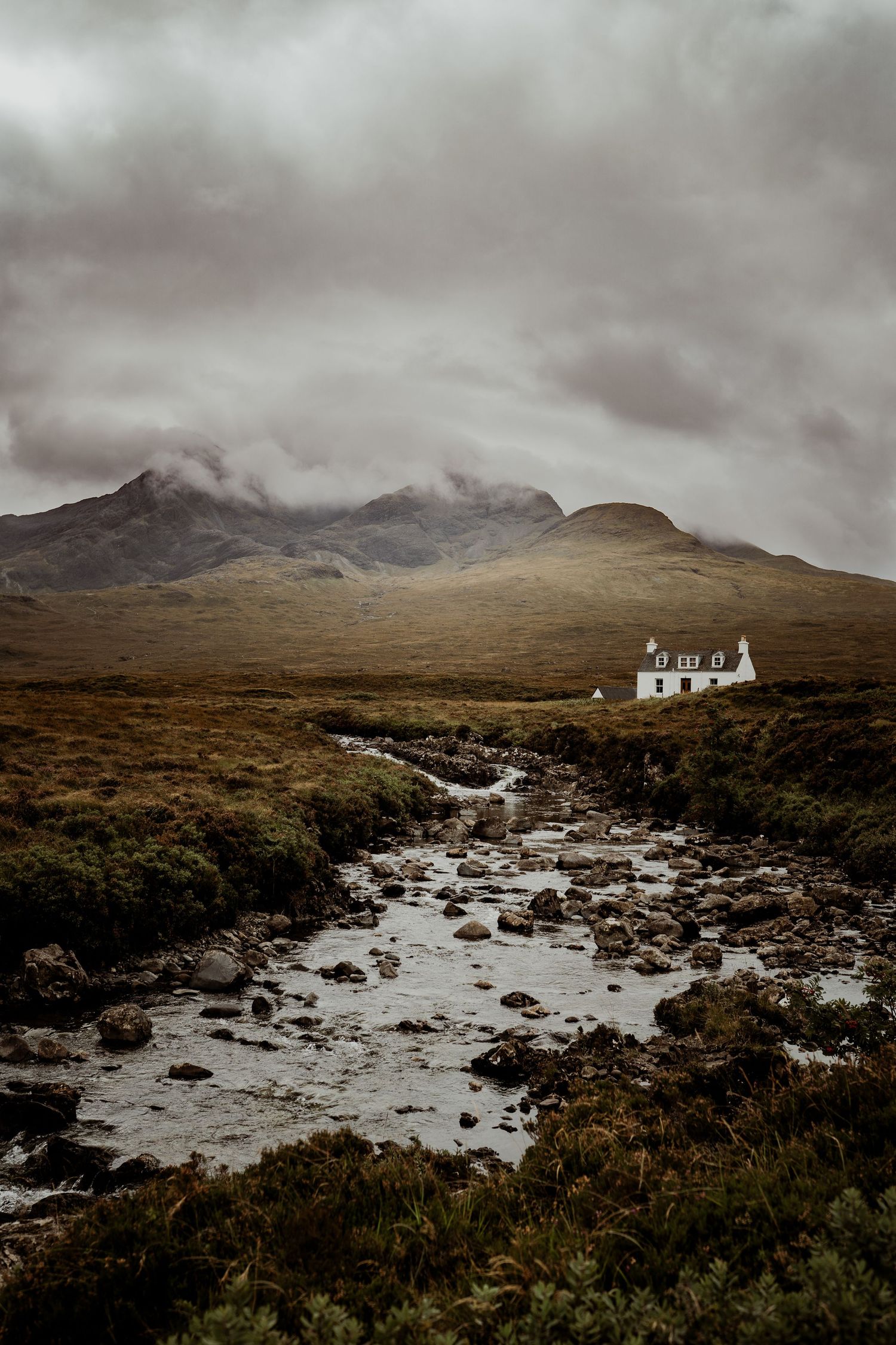 A white cottage sits beside a rocky stream in a moody Scottish Highland landscape with misty mountains.
