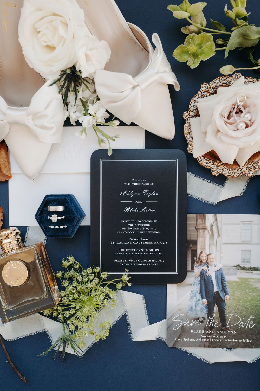 Elegant wedding flat lay with navy blue backdrop, black invitation suite, white roses, bridal heels, perfume, and save the date card.