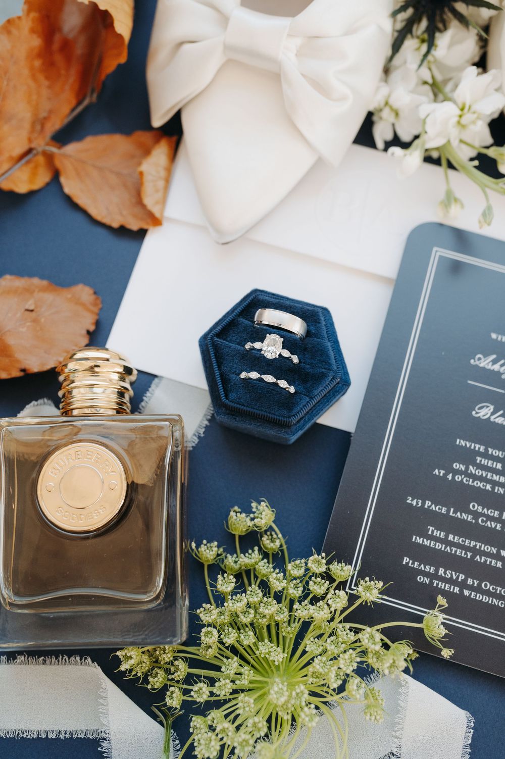 Navy blue wedding flat lay with velvet ring box, diamond rings, perfume bottle, floral invitation, bow tie, and autumn leaves.