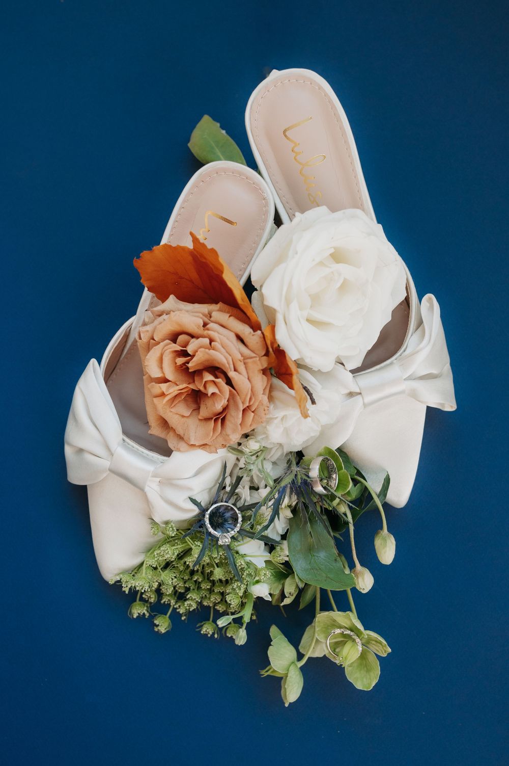 White bridal flats with bow detail rest on blue background beside ivory and rust roses, greenery, and a diamond engagement ring.