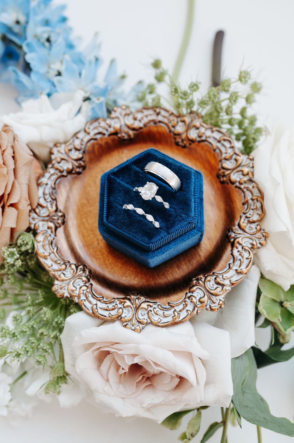 Wedding rings displayed in a blue velvet hexagon ring box on an ornate wooden tray surrounded by white and blush florals.